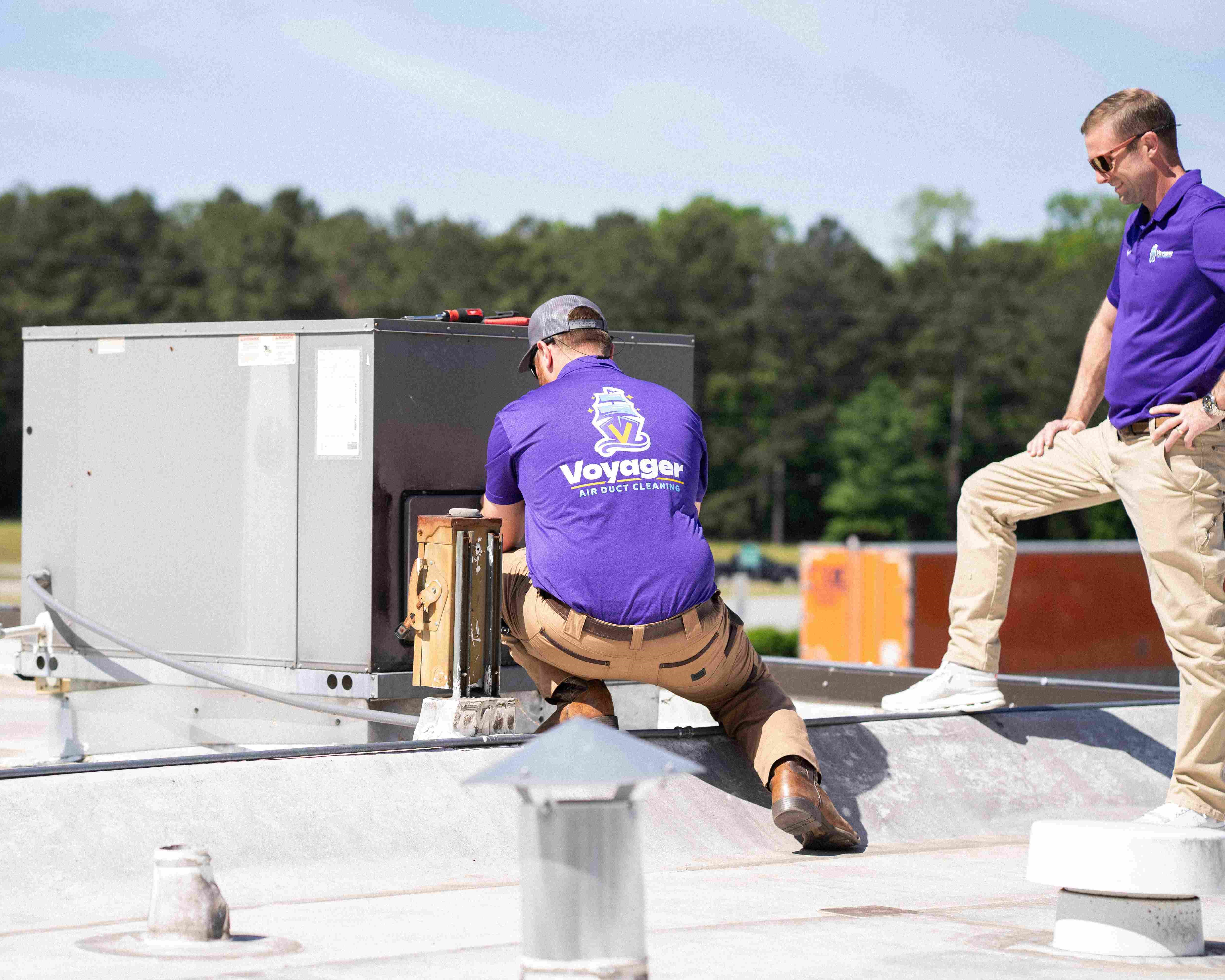 Two Voyager Air Duct Cleaning technicians servicing a commercial rooftop HVAC unit, one kneeling to access the unit while the other observes.